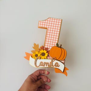 May include: A festive cake topper featuring a large number one with a peach and white gingham pattern. It is decorated with sunflowers, fall leaves, and an orange pumpkin. The banner reads "Camila" in gold glitter.