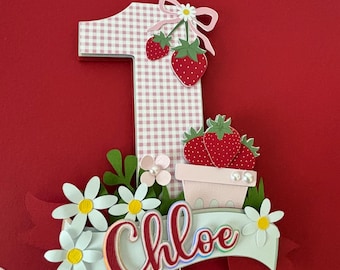 May include: A pink and white gingham number one cake topper with a strawberry design and the name "Chloe" on a blue banner. The topper is decorated with white daisies and green leaves.