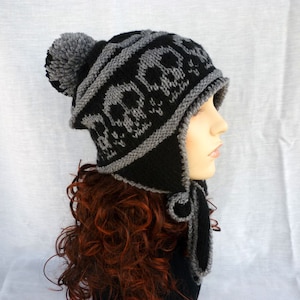 May include: Black and gray knitted hat with a skull pattern and a pom pom on top. The hat has ear flaps that tie under the chin.