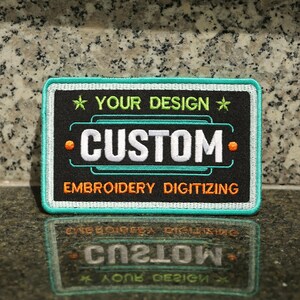 May include: Black embroidered patch with white text that says "CUSTOM" and orange text that says "EMBROIDERY DIGITIZING". The patch has a teal border and a white star with green text that says "YOUR DESIGN" above the word "CUSTOM".