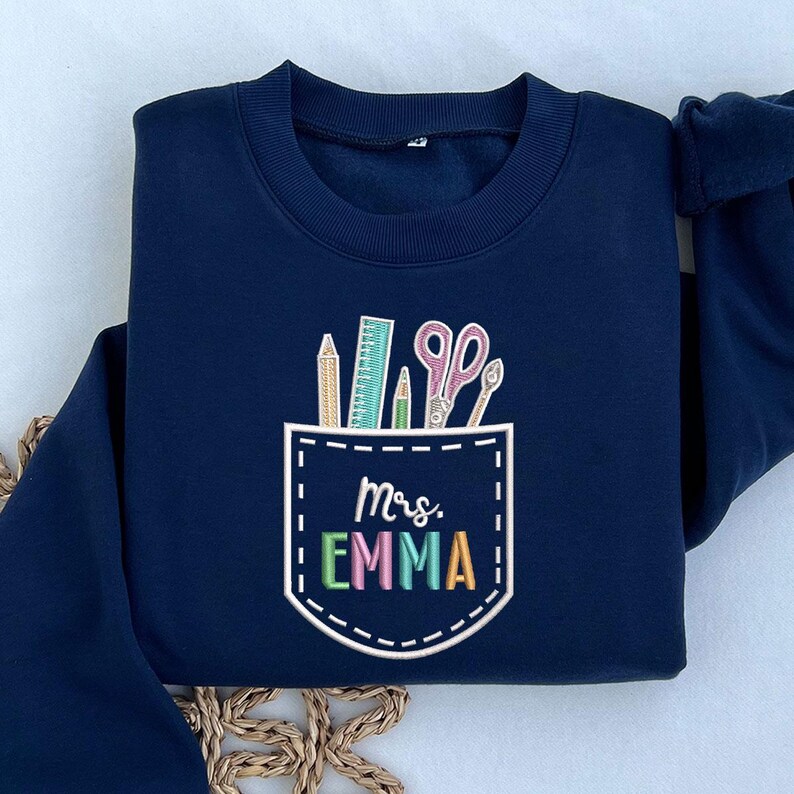 May include: Navy blue sweatshirt with a pocket design featuring school supplies. The pocket includes a pencil, ruler, scissors, and the text "Mrs. EMMA" in colorful letters. The sweatshirt is a crew neck style.
