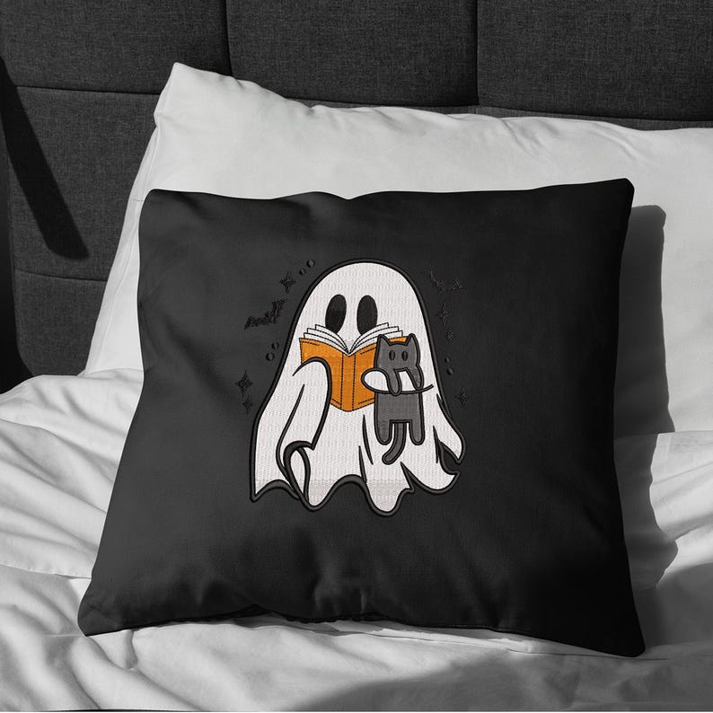 Puede incluir: Un coj&iacute;n rectangular negro con una ilustraci&oacute;n de un fantasma blanco. El fantasma sostiene un libro naranja abierto y un peque&ntilde;o gato gris. Peque&ntilde;os murci&eacute;lagos negros y destellos en forma de estrella rodean la figura central.