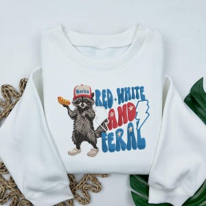 Op de afbeelding: Witte sweatshirt met een afbeelding van een wasbeer met een hoed met de tekst "Merica" en een hotdog. De tekst "Red, White, and Feral" is in rood, wit en blauw. Ronde hals.