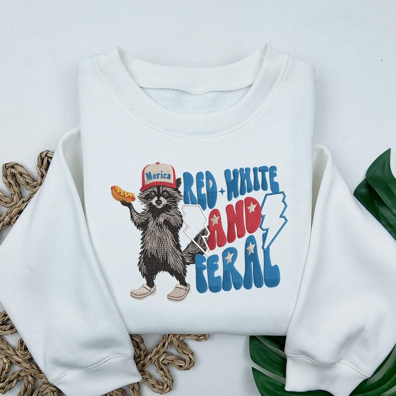 Op de afbeelding: Witte sweatshirt met een afbeelding van een wasbeer met een hoed met de tekst "Merica" en een hotdog. De tekst "Red, White, and Feral" is in rood, wit en blauw. Ronde hals.
