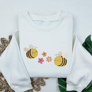 May include: White sweatshirt with embroidered bees and flowers. The bees are yellow and black with smiling faces. The flowers are pink and yellow.