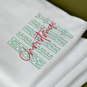 May include: White folded cloth with the words "MERRY Christmas" embroidered in green and red. The word "MERRY" is stacked vertically on the left side of the cloth, with "Christmas" written in a cursive font across the center.