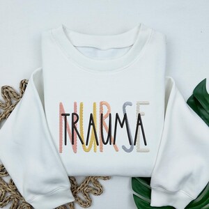 May include: White sweatshirt with the text "NURSE TRAUMA" embroidered in rainbow colors.