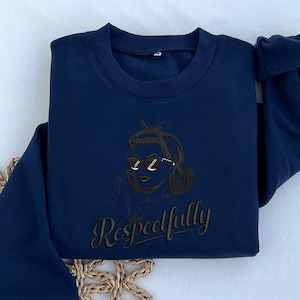May include: Navy blue sweatshirt featuring an embroidered design of a person wearing sunglasses and a headband. The word "Respectfully" is written in a cursive font below the design. The sweatshirt is folded on a light-colored surface.