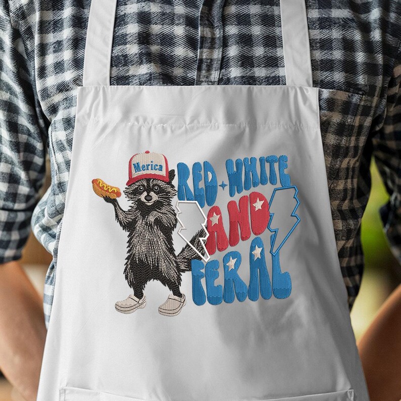 Op de afbeelding: Witte schort met een afbeelding van een wasbeer met een "Merica"-pet en een hotdog. De schort heeft de tekst "Red, White, and Feral" in rood en blauw. De wasbeer is zwart en wit.