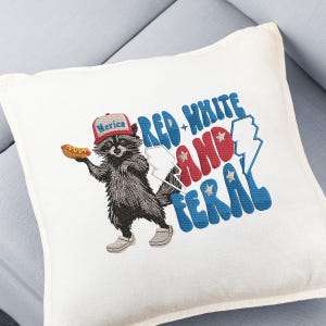 Op de afbeelding: Vierkant wit kussen met een geborduurd ontwerp. Het ontwerp toont een wasbeer met een hoed met de tekst "Merica" en een hotdog. De tekst luidt "Red, White, and Feral" in rood, wit en blauw.