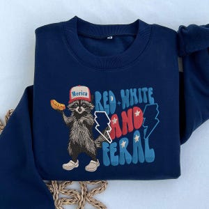 Op de afbeelding: Marineblauw sweatshirt met een geborduurde wasbeer met een "Merica" hoed en een hotdog. De tekst "Red White and Feral" is in rood, wit en blauw. Een leuk en patriottisch ontwerp.