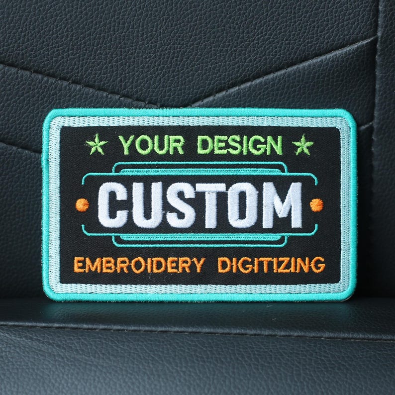 May include: A black and green embroidered patch with white text that reads "YOUR DESIGN" and "CUSTOM" with orange dots on either side of the word "CUSTOM". The text "EMBROIDERY DIGITIZING" is below the word "CUSTOM".