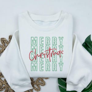 May include: White sweatshirt with the words "MERRY" stacked vertically in green, with "Christmas" in red script overlaid. The sweatshirt has a crew neck and long sleeves, perfect for the holiday season.