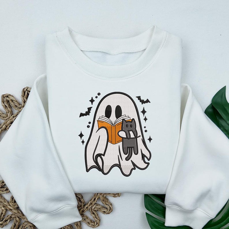 Puede incluir: Una sudadera blanca de cuello redondo con un gr&aacute;fico central de un fantasma leyendo un libro naranja mientras sostiene un peque&ntilde;o gato negro. El dise&ntilde;o est&aacute; decorado con peque&ntilde;os murci&eacute;lagos negros y estrellas de cuatro puntas.