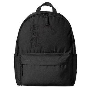 May include: Black backpack with a top handle, front pocket, and a main compartment with a zipper closure. The backpack features the phrase "HEY, YOU DROPPED THIS" and a portrait of a person.