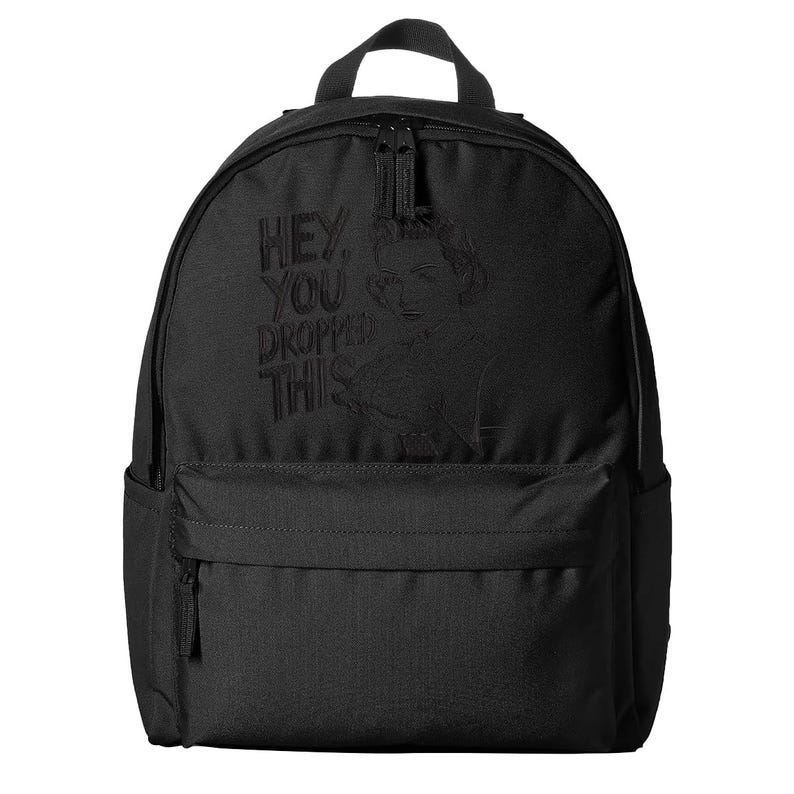 May include: Black backpack with a top handle, front pocket, and a main compartment with a zipper closure. The backpack features the phrase "HEY, YOU DROPPED THIS" and a portrait of a person.