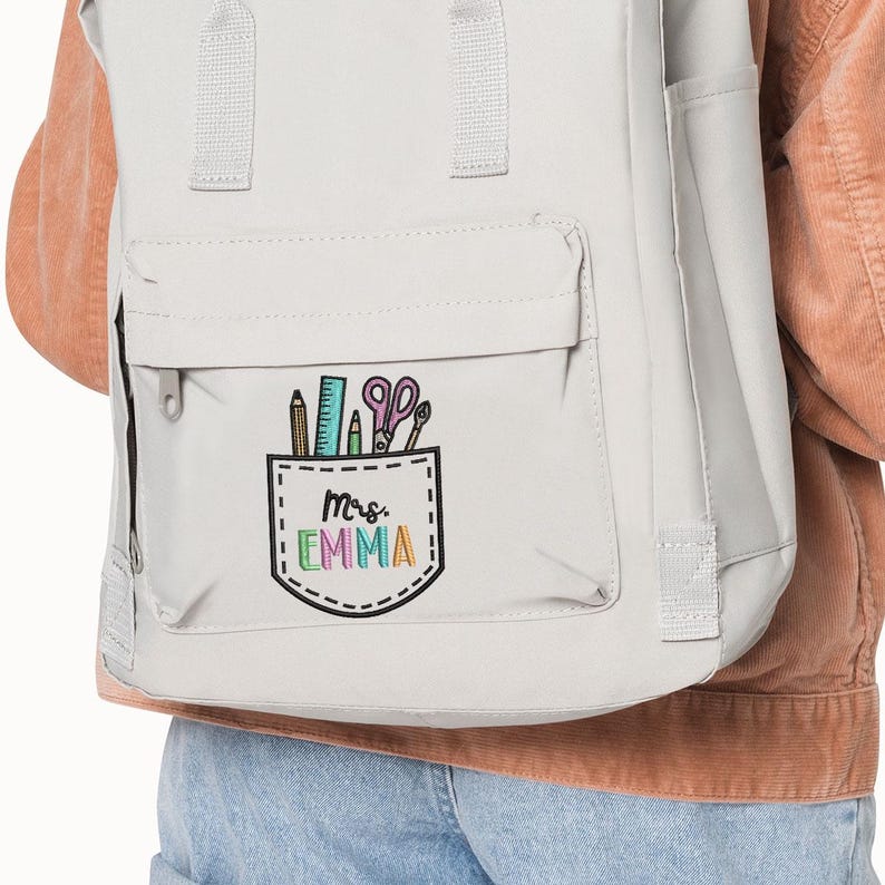 May include: A light gray backpack with a front pocket featuring a colorful embroidered design of school supplies and the name "Mrs. Emma". The design includes a ruler, scissors, and pencils.
