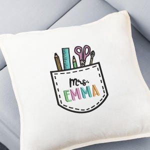 May include: A white square pillow with an embroidered pocket design. The pocket contains a pencil, ruler, pen, scissors, and a small tool. The text "Mrs. EMMA" is embroidered in colorful letters below the pocket.