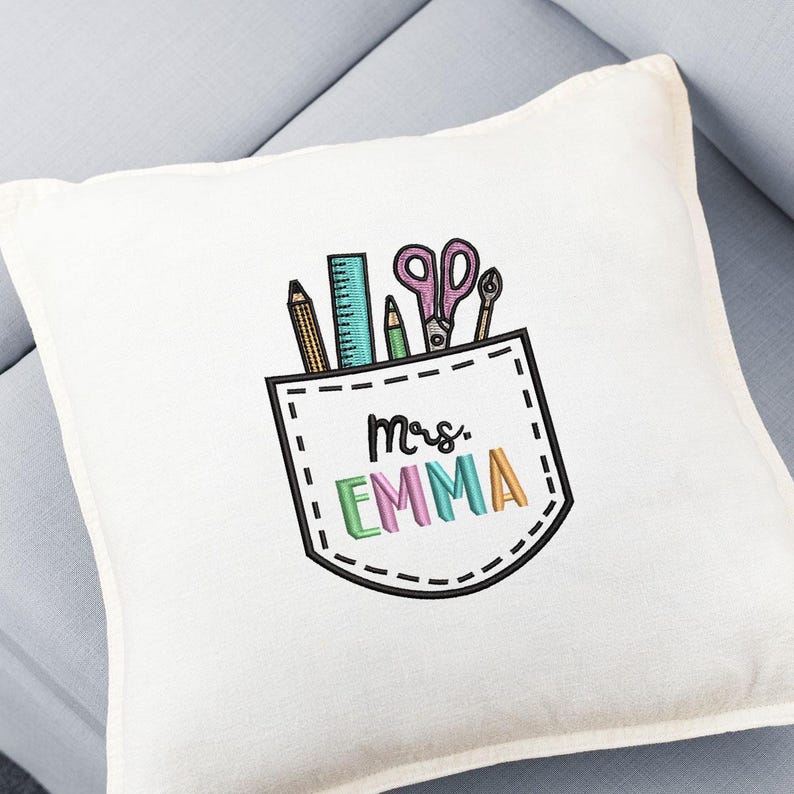 May include: A white square pillow with an embroidered pocket design. The pocket contains a pencil, ruler, pen, scissors, and a small tool. The text "Mrs. EMMA" is embroidered in colorful letters below the pocket.