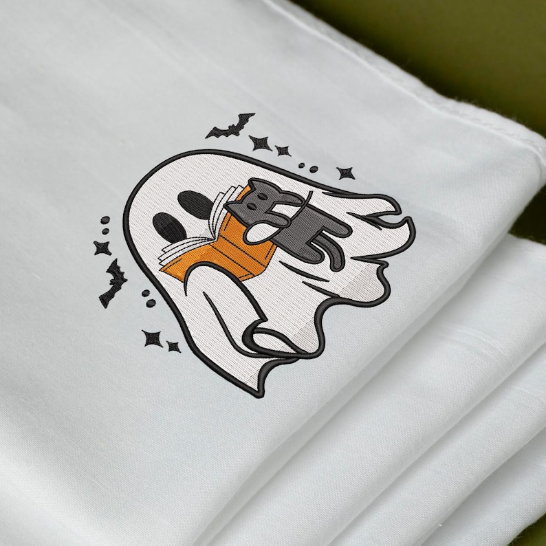Puede incluir: Un art&iacute;culo de tela blanca con un dise&ntilde;o bordado de un fantasma blanco que sostiene un libro naranja. Un peque&ntilde;o gato negro est&aacute; posado sobre el libro. El dise&ntilde;o est&aacute; rodeado de peque&ntilde;os murci&eacute;lagos negros bordados y estrellas de cuatro puntas.