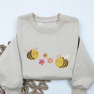 May include: A beige sweatshirt with embroidered bees and flowers. The bees are yellow and black with smiling faces. The flowers are pink, yellow, and orange.