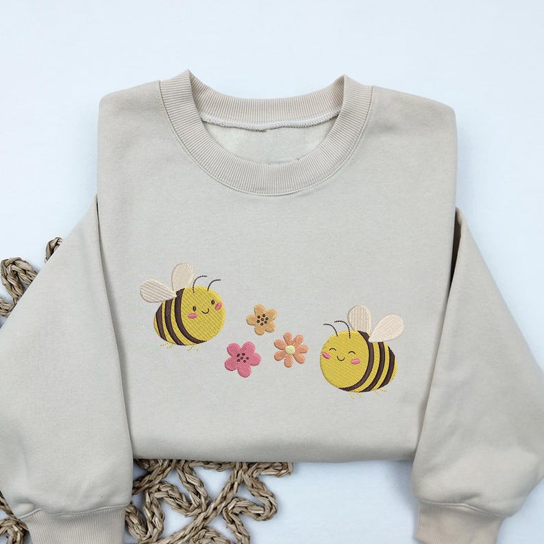 May include: A beige sweatshirt with embroidered bees and flowers. The bees are yellow and black with smiling faces. The flowers are pink, yellow, and orange.