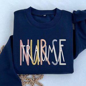 May include: A navy blue sweatshirt with the words "NURSE" and "TRAUMA" embroidered in colorful thread.
