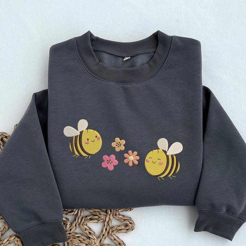 May include: A dark gray sweatshirt with a cute embroidered design of two smiling bees and flowers. The bees are yellow and black with white wings and the flowers are pink, yellow, and orange.