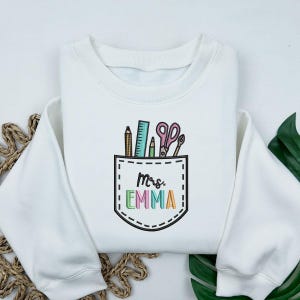 May include: White sweatshirt with a pocket design featuring school supplies: pencils, a ruler, scissors, and a pen. The pocket has the name "Mrs. EMMA" embroidered in colorful letters. A cozy, casual top.