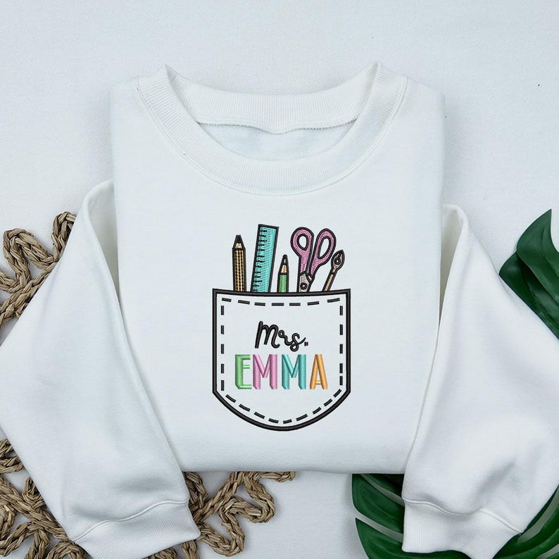 May include: White sweatshirt with a pocket design featuring school supplies: pencils, a ruler, scissors, and a pen. The pocket has the name "Mrs. EMMA" embroidered in colorful letters. A cozy, casual top.