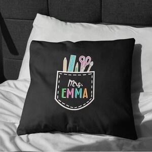 May include: Black decorative pillow with a pocket design featuring school supplies and the text "Mrs. Emma" in colorful letters. The pocket design includes a pencil, ruler, scissors, and other school supplies.