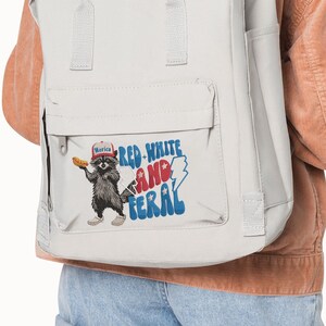 Op de afbeelding: Een lichtgrijze rugzak met een voorvak met een afbeelding van een wasbeer met een hotdog en de tekst "Red, White, and Feral". De wasbeer draagt een hoed met de tekst "Merica".