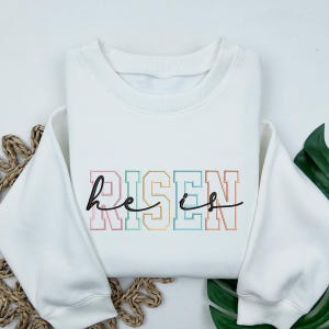May include: White sweatshirt with a rainbow colored graphic that says "He is Risen" in a varsity style font.