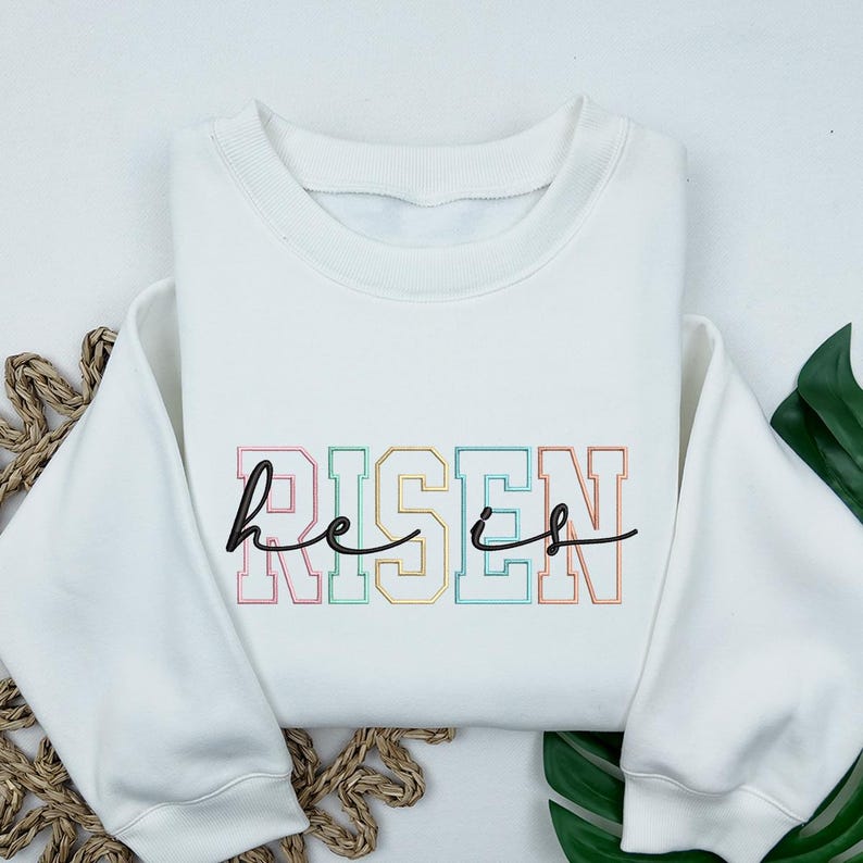 May include: White sweatshirt with a rainbow colored graphic that says "He is Risen" in a varsity style font.
