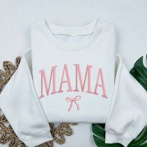May include: White sweatshirt with the word "MAMA" embroidered in pink with a pink bow underneath.