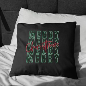 May include: Black square pillow with the words "MERRY CHRISTMAS" in a repeating pattern. The word "MERRY" is in a green outline font, and "Christmas" is in a red script font. The pillow is on a white bed.