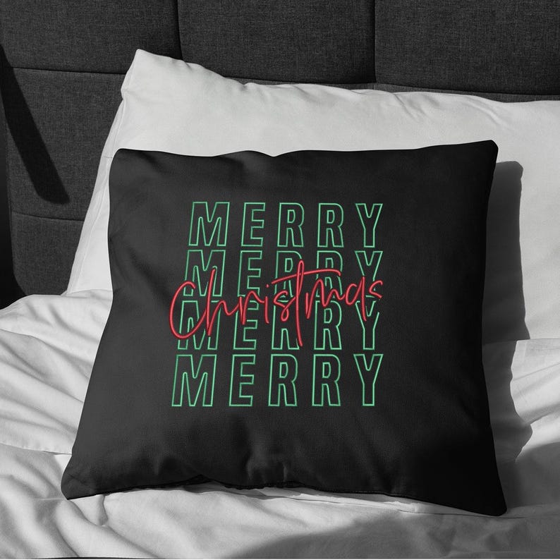 May include: Black square pillow with the words "MERRY CHRISTMAS" in a repeating pattern. The word "MERRY" is in a green outline font, and "Christmas" is in a red script font. The pillow is on a white bed.