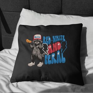 Op de afbeelding: Zwart kussen met een wasbeer die een hoed draagt met "Merica" erop en een hotdog vasthoudt. De tekst "Red, White, and Feral" is in rood en blauw. De wasbeer draagt witte schoenen. Een leuk, patriottisch ontwerp.