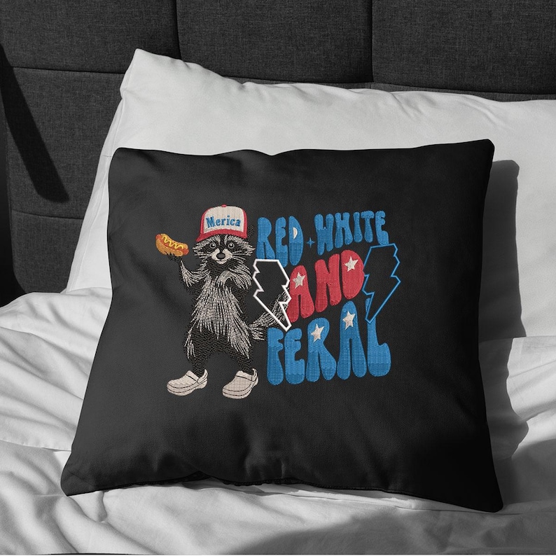 Op de afbeelding: Zwart kussen met een wasbeer die een hoed draagt met "Merica" erop en een hotdog vasthoudt. De tekst "Red, White, and Feral" is in rood en blauw. De wasbeer draagt witte schoenen. Een leuk, patriottisch ontwerp.
