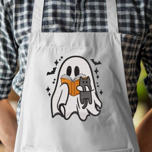 Puede incluir: Un delantal blanco con un gr&aacute;fico central de un fantasma blanco leyendo un libro naranja mientras sostiene un peque&ntilde;o gato gris. El dise&ntilde;o incluye contornos negros, peque&ntilde;os murci&eacute;lagos negros y detalles en forma de estrella.