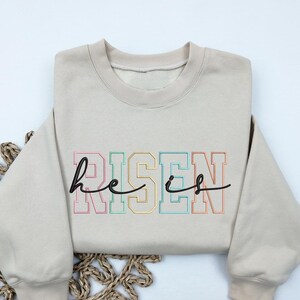 May include: A beige sweatshirt with the text "he is risen" in black lettering with rainbow colored outlines.