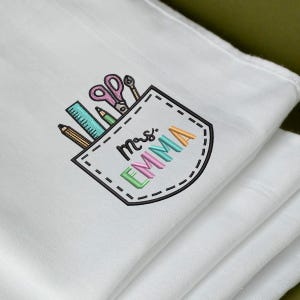 May include: White cloth with an embroidered pocket design featuring school supplies. The pocket includes a ruler, pencil, scissors, and the name "Mrs. EMMA" in colorful letters. The design is stitched in various colors, including pink, green, and orange.