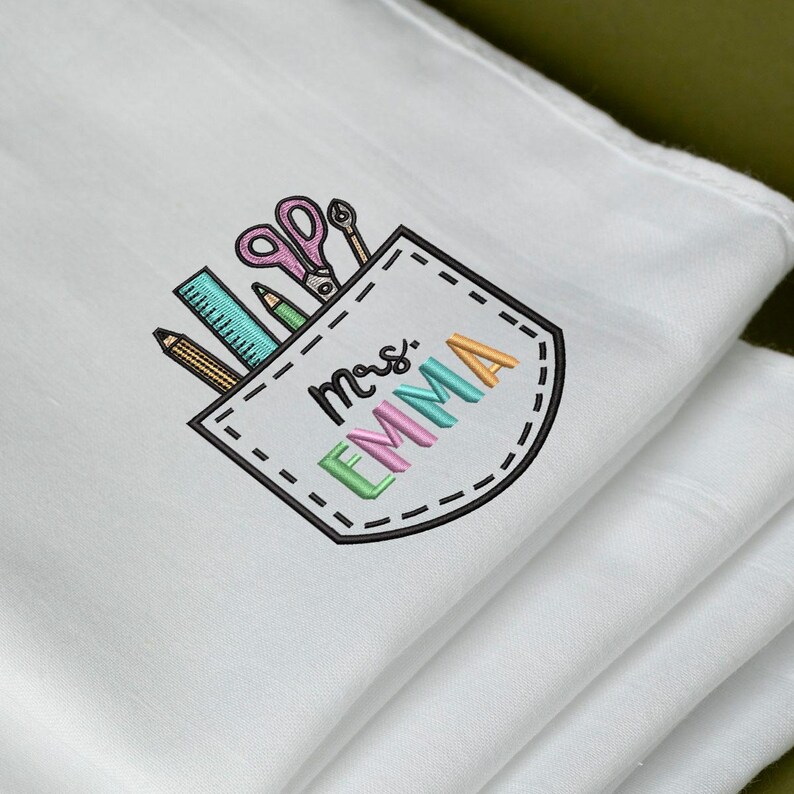 May include: White cloth with an embroidered pocket design featuring school supplies. The pocket includes a ruler, pencil, scissors, and the name "Mrs. EMMA" in colorful letters. The design is stitched in various colors, including pink, green, and orange.