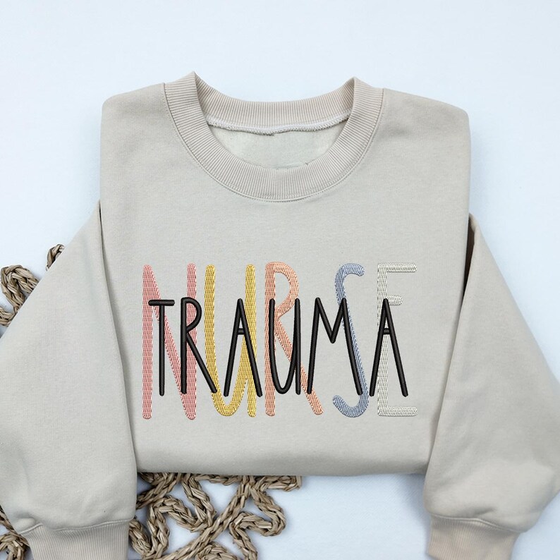 May include: A beige sweatshirt with the word "TRAUMA" embroidered in black letters with rainbow colored outlines.