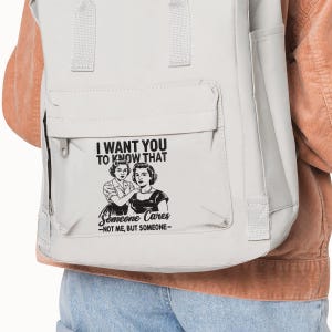 May include: A light gray backpack with a flap closure and a front pocket. The backpack features a graphic with the text "I WANT YOU TO KNOW THAT" above an illustration of two women and the text "Someone Cares -NOT ME, BUT SOMEONE-".