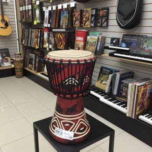 May include: A red djembe drum with a natural drumhead and black rope accents. The drum features white geometric designs and is displayed on a black stand in a music shop. Musical instruments and books are visible in the background.