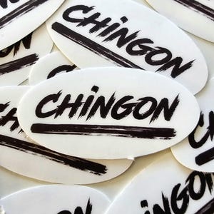 May include: A close-up of several oval-shaped stickers with the word "CHINGON" written in black brushstrokes on a white background.