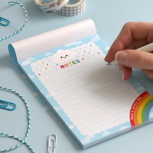 May include: A blue notepad with a white cloud and raindrops design. The notepad has the word "NOTES" written in rainbow letters. The bottom right corner of the notepad has a rainbow with the text "have a lovely day".