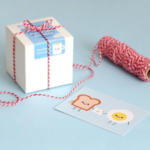 May include: A white gift box tied with red and white baker's twine. The box has a label that says "handmade doodle" and "Handkerchief Pincushion". A card with a cartoon illustration of a slice of bread and an egg with the text "bc" is next to the box.
