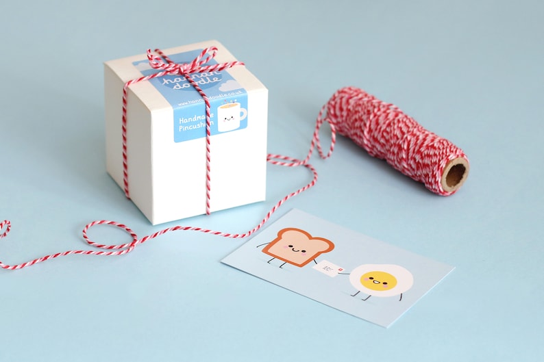 May include: A white gift box tied with red and white baker's twine. The box has a label that says "handmade doodle" and "Handkerchief Pincushion". A card with a cartoon illustration of a slice of bread and an egg with the text "bc" is next to the box.